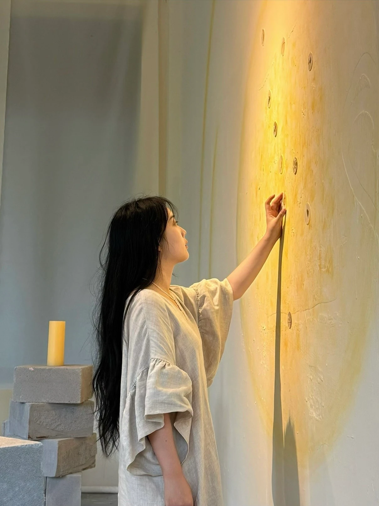 Na Xin reaching toward small circular forms on a yellow-lit wall, beside stacked blocks.