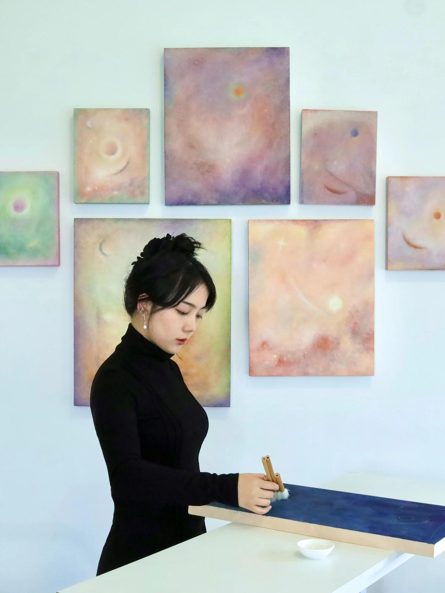 Na Xin working with brushes on a blue wood panel, with paintings displayed behind her.